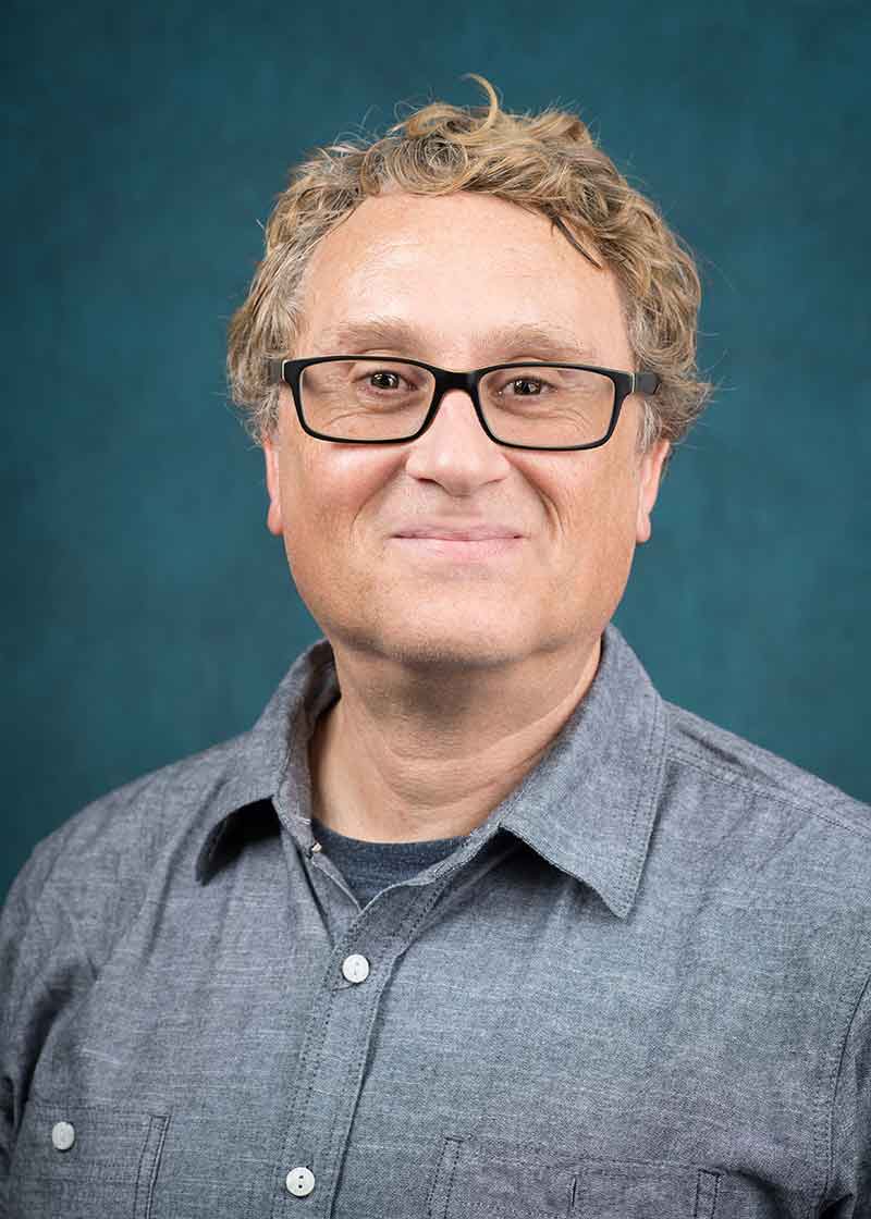 Headshot of a man with black-rimmed glasses and a grey button-down dress shirt