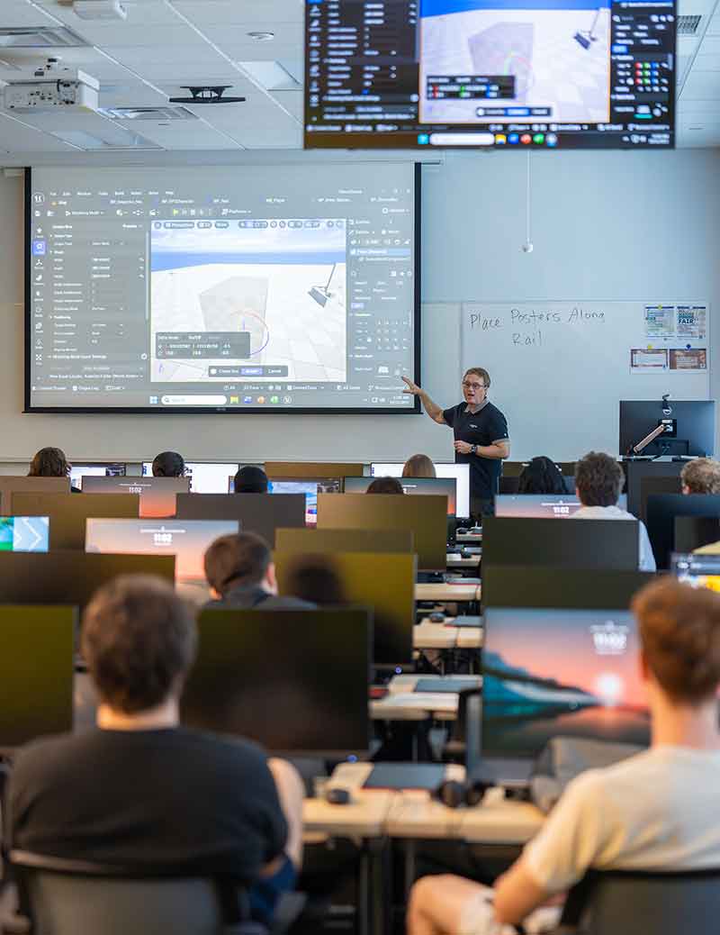 Classroom with students at computer stations watching a large screen showing 3D design software