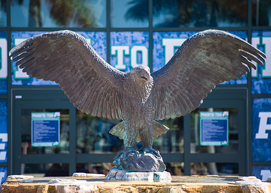 Bronze eagle statue with wings fully spread, mounted on a stone pedestal in front of a building entrance with large glass doors.