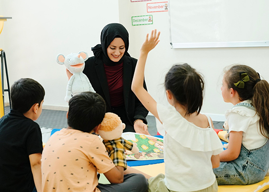 A teacher sits on the floor with four children in a classroom, holding a white mouse puppet. One child raises a hand while others listen attentively. The floor has a colorful mat, and posters labeled “November” and “December” are visible on the wall behind them.