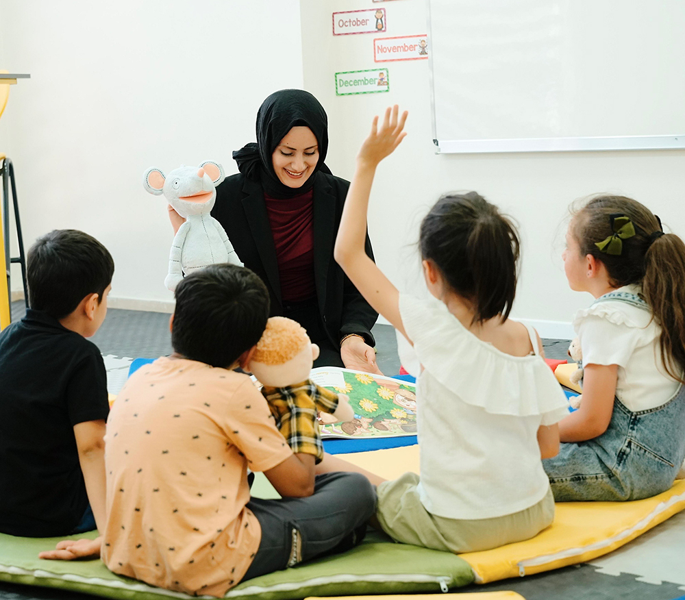 A teacher sits on the floor with four children in a classroom, holding a white mouse puppet. One child raises a hand while others listen attentively. The floor has a colorful mat, and posters labeled “November” and “December” are visible on the wall behind them.