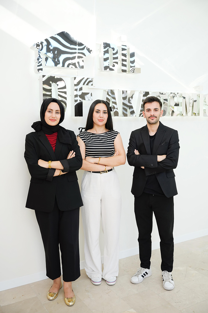 Three individuals stand side by side with arms crossed in front of a white wall displaying the words “DRAK ACADEMY.” The person on the left wears a black suit with a red top and gold shoes; the middle person wears a black and white striped top with white pants and shoes; the person on the right wears a black suit with a black shirt and white sneakers.