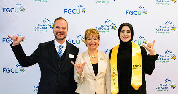 Three people stand in front of a backdrop with the Florida Gulf Coast University (FGCU) logo. The person on the left is wearing a dark suit and tie, the person in the middle is wearing a light-colored blazer, and the person on the right is wearing a black outfit with a yellow honors stole that has "FGCU" and "HONORS" written on it.