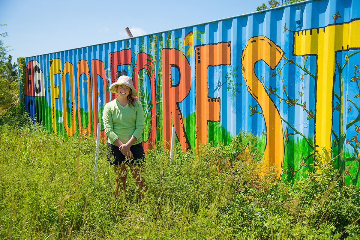 A person wearing a wide-brimmed hat and light green long-sleeve shirt sits in tall grass in front of a brightly painted shipping container. The container features large, colorful letters spelling 'FOOD FOREST' with plant and vine illustrations. The sky is clear and blue, indicating a sunny day.