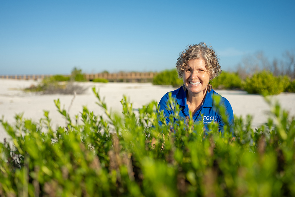 Person in FGCU shirt standing behind green shrubs on a sandy beach with clear blue sky.