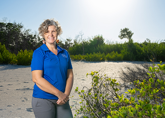 Person in a blue polo shirt standing on a sandy beach with green shrubs and trees in the background.
