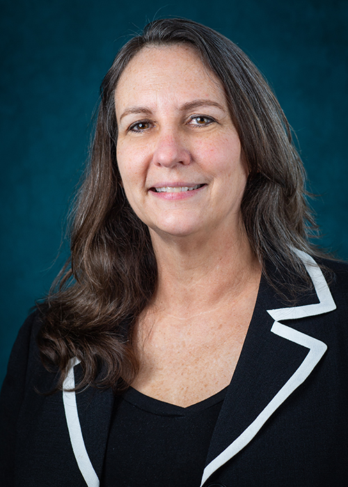 A person with long, wavy hair is shown from the shoulders up against a dark teal background. They are wearing a black blazer with white trim and a black top, looking directly at the camera with a neutral expression
