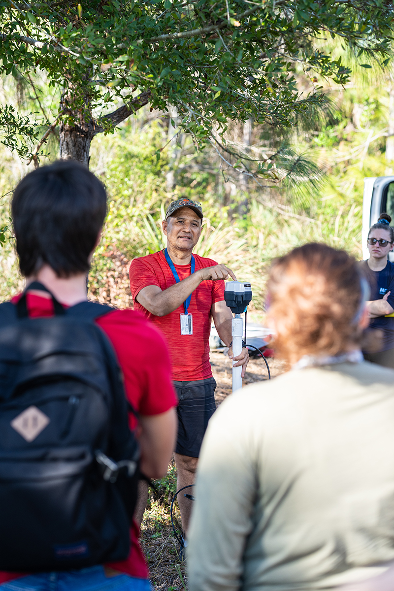 A person wearing a red shirt, black shorts, cap, and ID badge stands under a tree holding equipment and speaking to a small group of people with backpacks. The group is outdoors in a sunlit, wooded area with greenery in the background