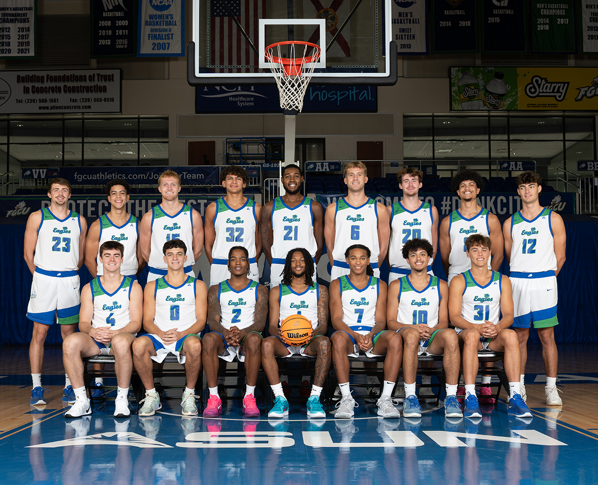 Group photo of a basketball team in a gymnasium, with players wearing white and blue uniforms that feature green accents and the word “Eagles” on the front. The team is arranged in two rows: eight players seated in the front row and seven players standing behind them. A basketball is held by a player in the center of the front row. A basketball hoop and banners are visible in the background.