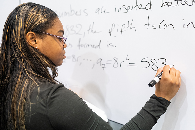 Person writing mathematical expressions and partial sentences on a whiteboard using a black marker, including exponential notation and sequences.