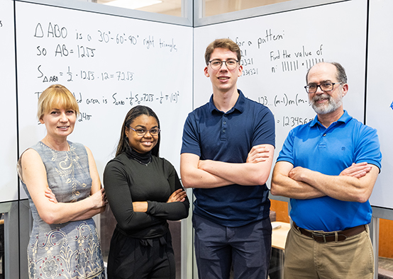 Four individuals stand in front of a whiteboard filled with mathematical equations and geometry problems. The group is posed with arms crossed, and the whiteboard displays triangle area calculations, algebraic expressions, and number theory formulas.