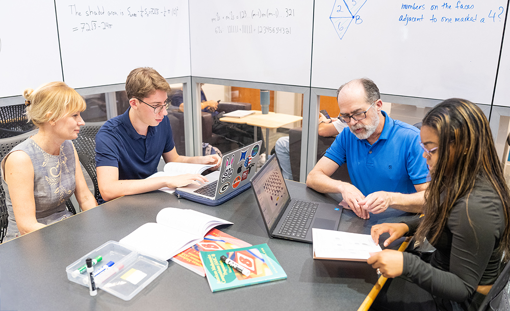 Four individuals sit around a table in a study room, discussing math problems with open books and laptops. A whiteboard behind them displays equations, including algebraic expressions and a homological algebra reference. Books titled “Calculus” and “Algebra” are visible on the table.