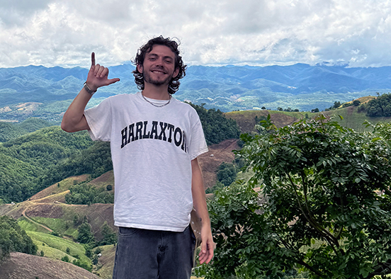 A person stands on a hillside with rolling green hills and distant mountains in the background. They wear a white "HARLAXTON" t-shirt and dark pants, with curly hair,