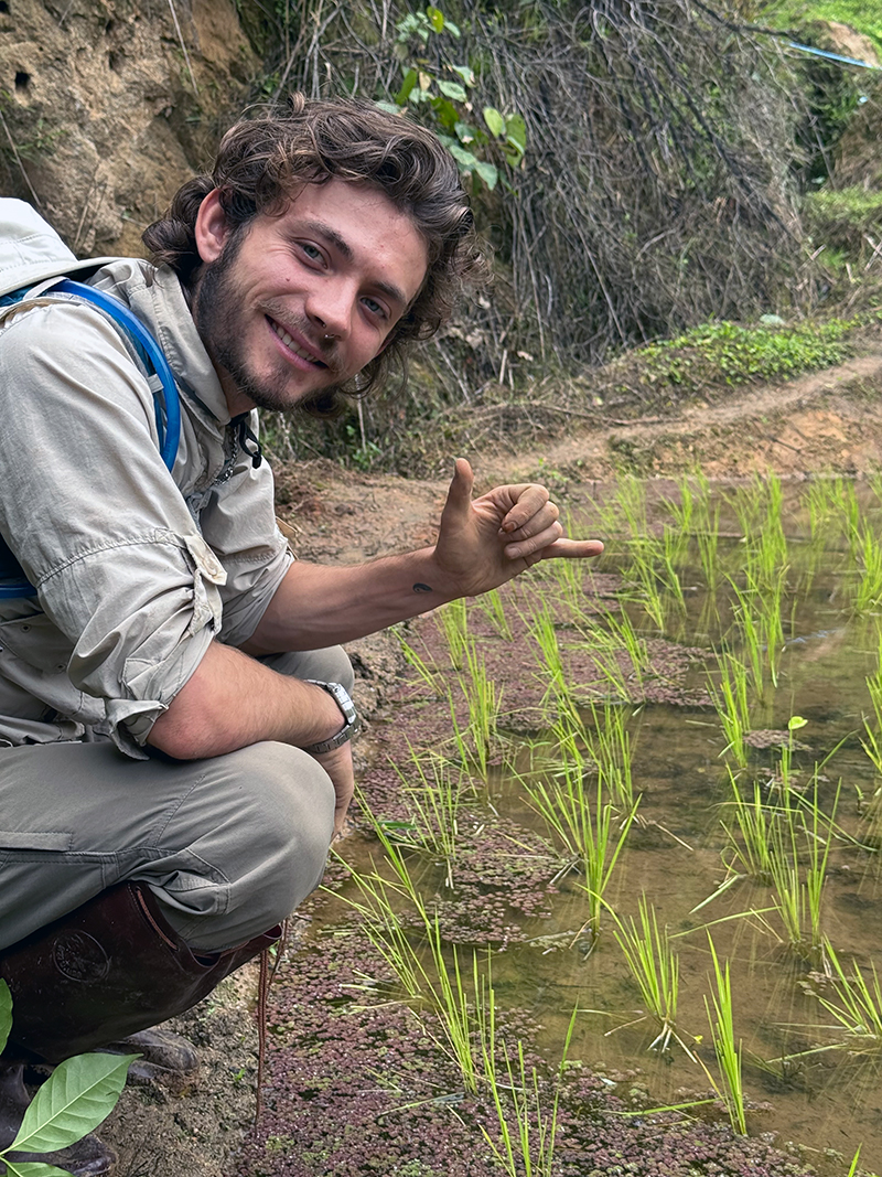 A person in outdoor clothing crouches beside a small body of water with young green plants growing in it. They wear a backpack and make a hand gesture with the thumb and pinky finger extended. The surrounding area includes earthy terrain, rocks, and scattered greenery.