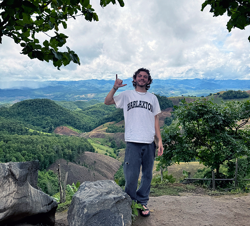 A person stands on a hillside with rolling green hills and distant mountains in the background. They wear a white "HARLAXTON" t-shirt and dark pants, with curly hair,