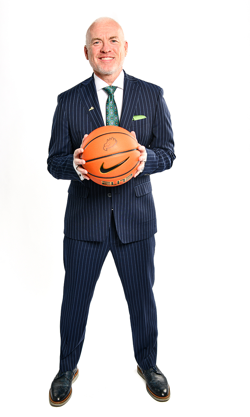 Person wearing a dark blue pinstripe suit with a white dress shirt, green patterned tie, and green pocket square, standing and holding an orange Nike basketball at chest level against a plain white background.