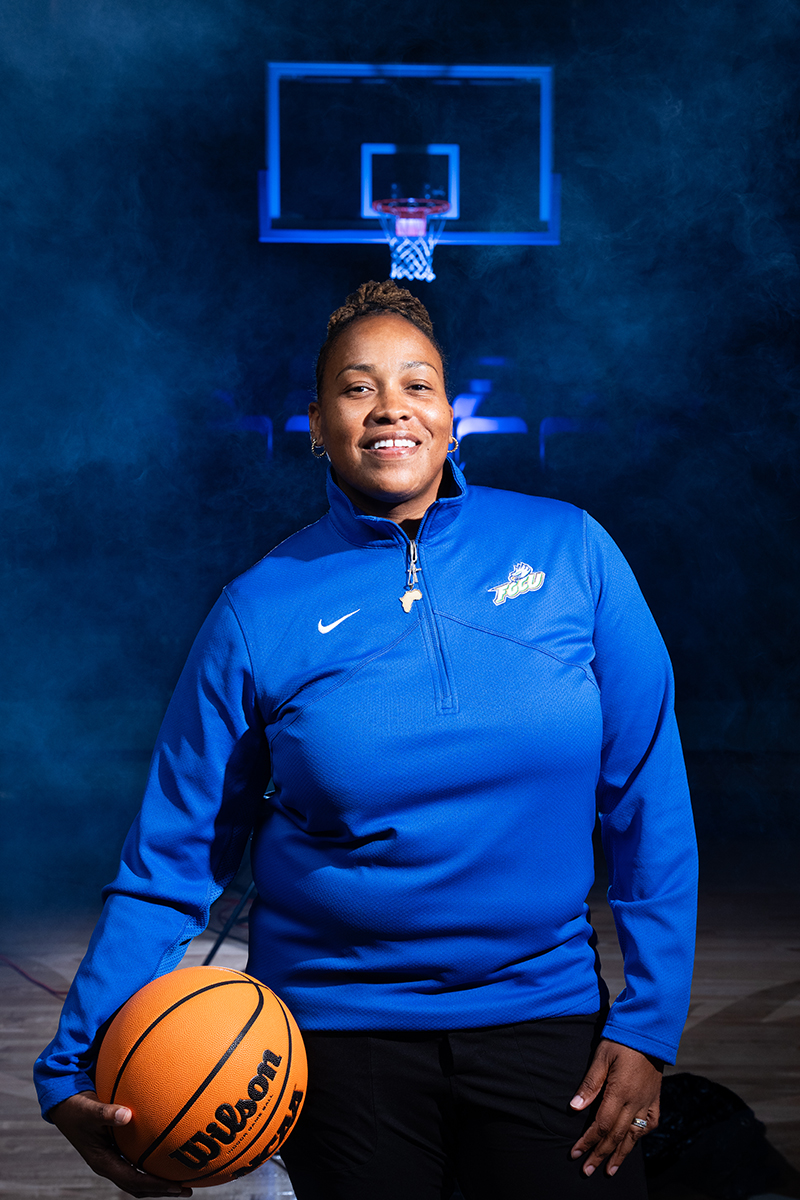 Person wearing a royal blue athletic pullover with an embroidered logo on the left chest, standing on a basketball court with a basketball held in one hand. A basketball hoop is visible in the background with dramatic blue lighting and mist creating a bold, high-contrast atmosphere.