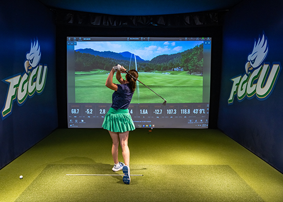 Person mid-swing in an indoor golf simulator at Florida Gulf Coast University, facing a screen displaying a virtual course and swing metrics like speed, launch angle, and spin rate. FGCU eagle logos are visible on the walls