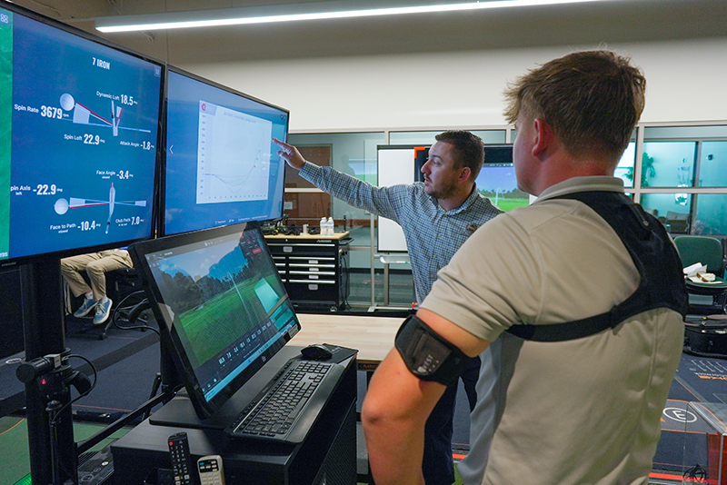 Two individuals in a golf swing analysis lab review data on multiple monitors. One person wears a harness and arm sensor while observing swing metrics like spin rate and launch angle displayed on screens.