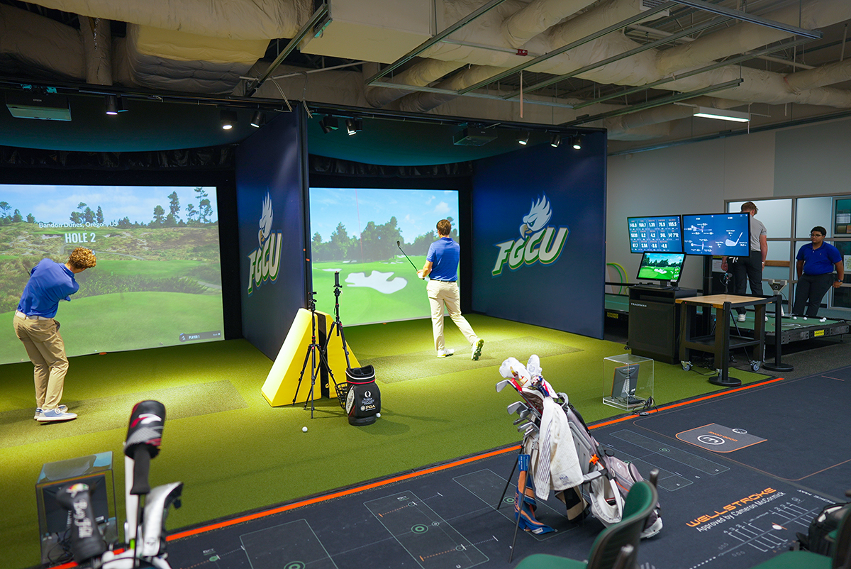 Indoor golf simulation room at Florida Gulf Coast University with two people practicing swings in front of virtual course screens, golf bags on turf, and others using performance analysis equipment. FGCU logos are visible on the walls.