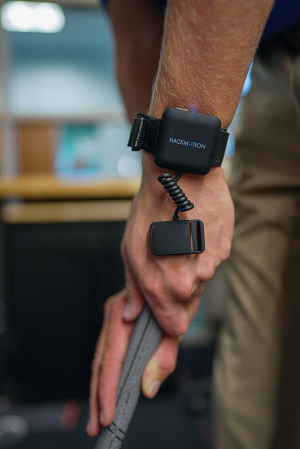Close-up of a person gripping a golf club indoors, wearing a black HackMotion wristband with a connected motion-tracking device on the hand.