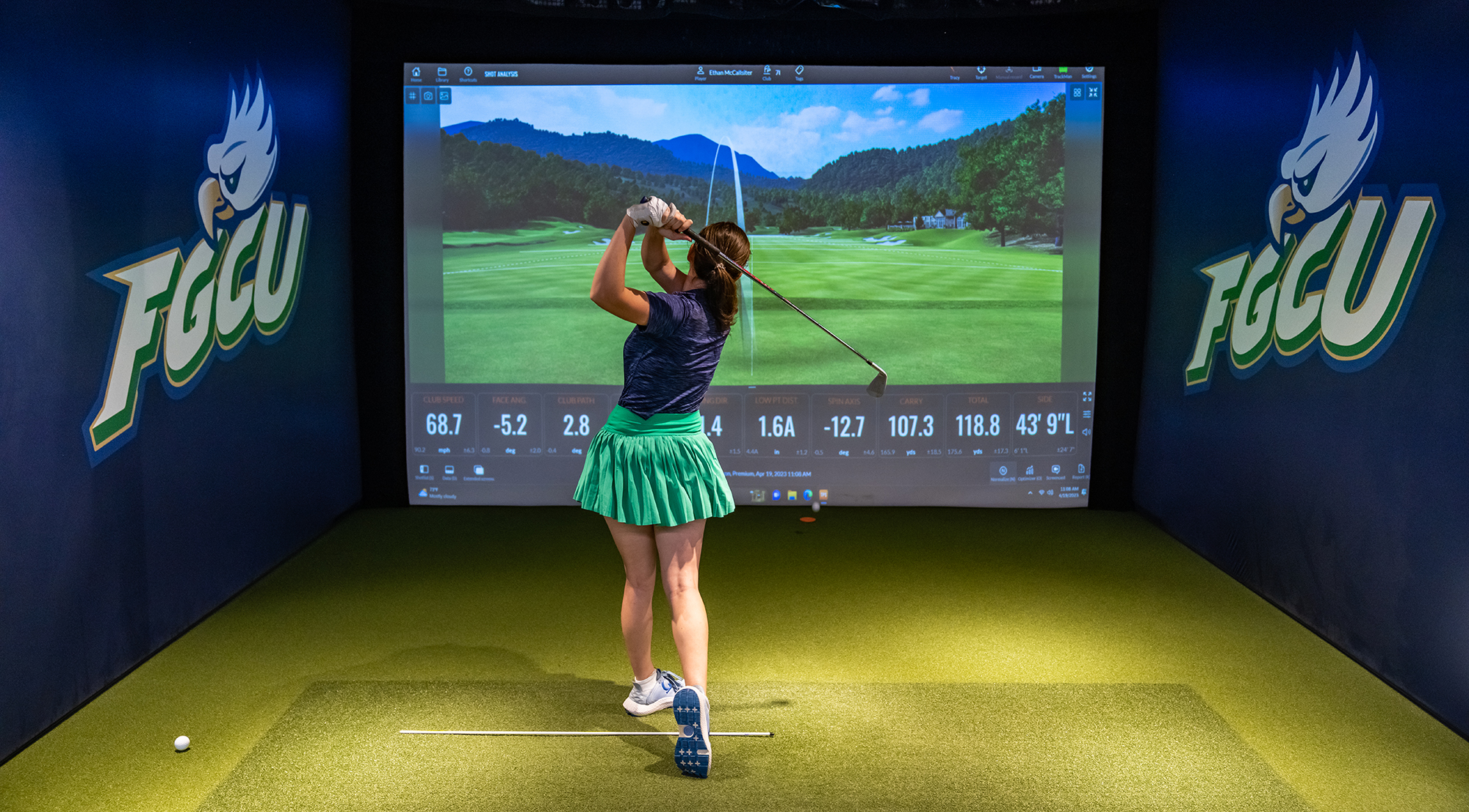 Person mid-swing in an indoor golf simulator at Florida Gulf Coast University, facing a screen displaying a virtual course and swing metrics like speed, launch angle, and spin rate. FGCU eagle logos are visible on the walls