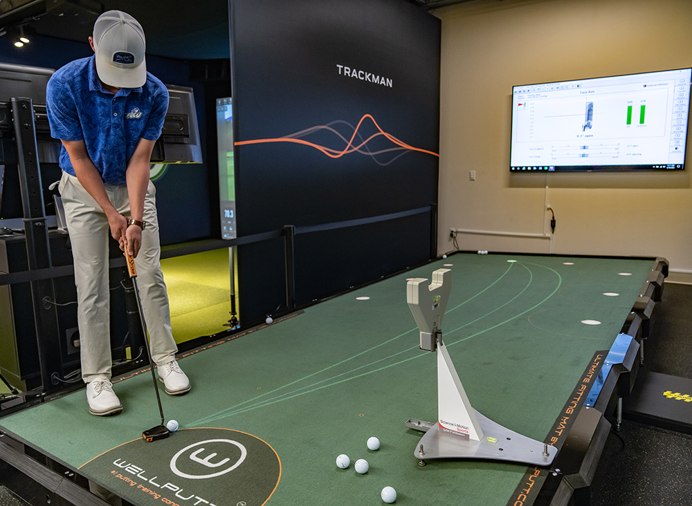 Person practicing putting on an indoor Wellputt golf green with scattered golf balls, a data-tracking device near the hole, and a screen displaying performance metrics.