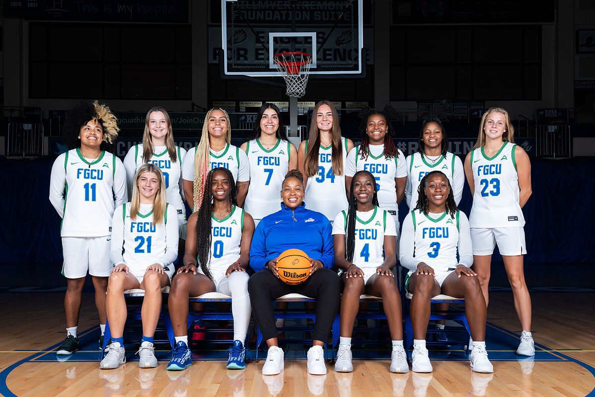 Group photo of a basketball team in a gymnasium, featuring players wearing white FGCU uniforms with green and blue accents. The team is arranged in two rows: eight players seated in the front row and seven players standing behind them. A person in the center of the front row wears a bright blue athletic pullover and holds an orange basketball. A basketball hoop is visible in the background above the group.