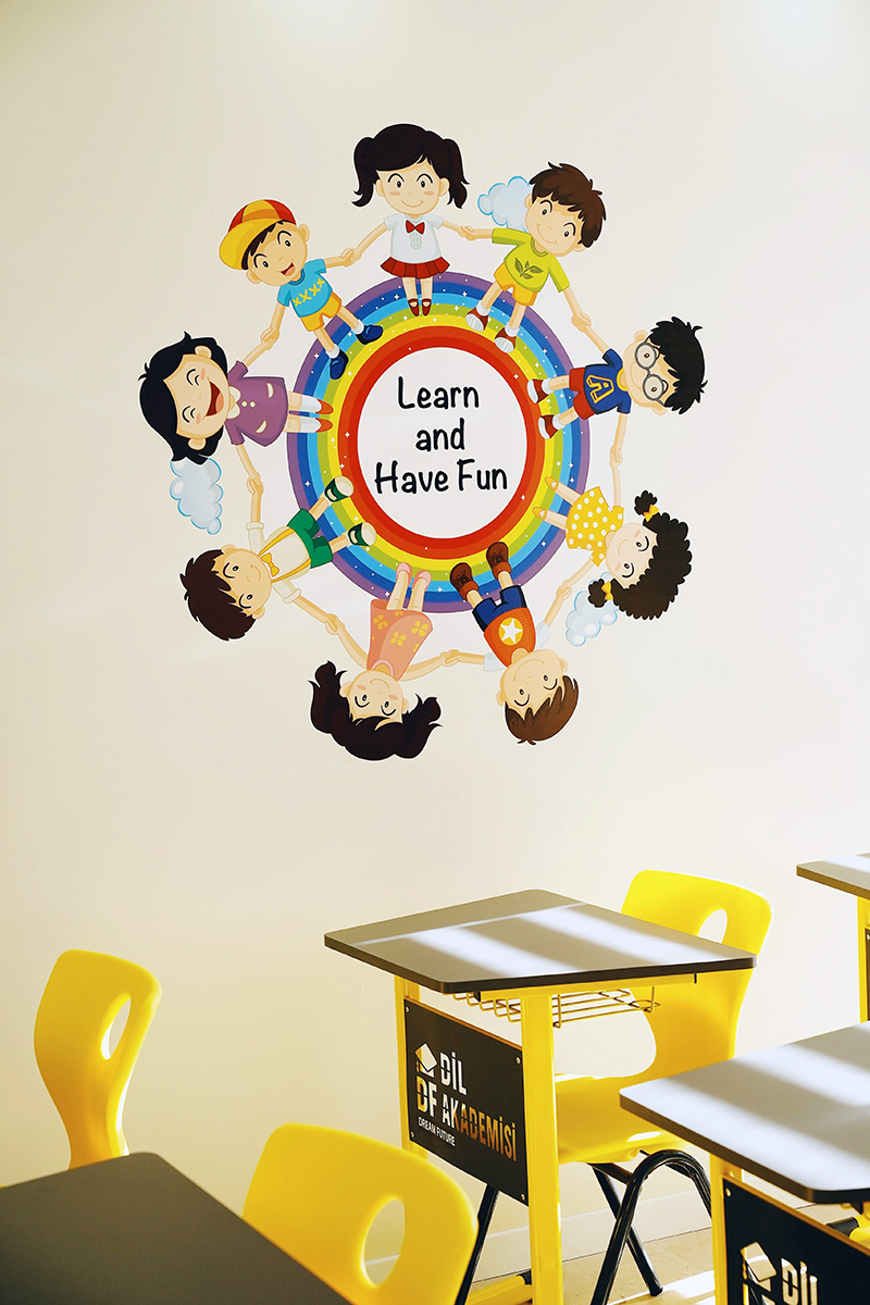 A classroom wall displays a colorful mural of eight cartoon children holding hands around a red-bordered white circle with the words “Learn and Have Fun.” The children wear bright outfits and appear to be smiling. Below the mural are yellow chairs and black-topped desks.