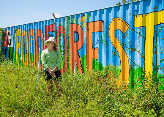 A person wearing a wide-brimmed hat and light green long-sleeve shirt sits in tall grass in front of a brightly painted shipping container. The container features large, colorful letters spelling 'FOOD FOREST' with plant and vine illustrations. The sky is clear and blue, indicating a sunny day.