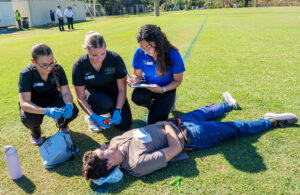 Medical team kneels on grass assessing a person lying down during emergency drill.