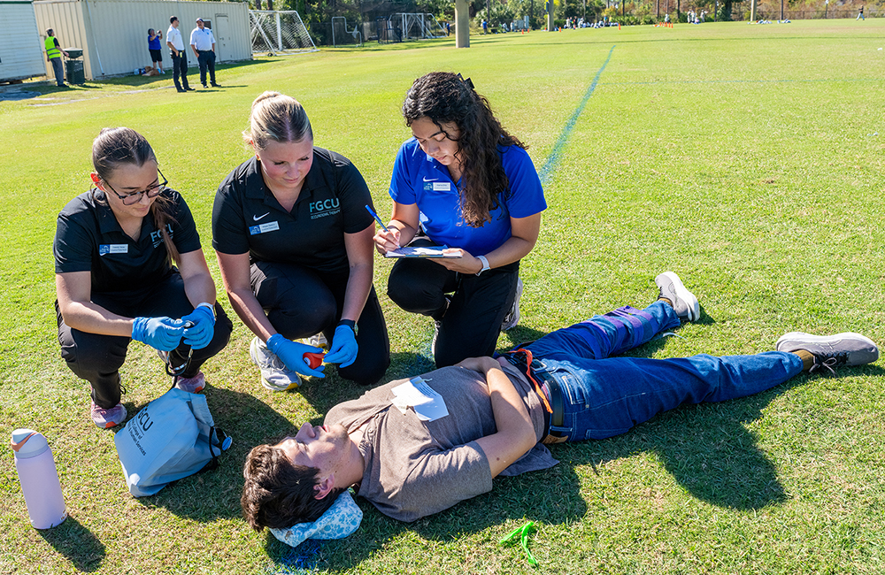 Medical team kneels on grass assessing a person lying down during emergency drill.