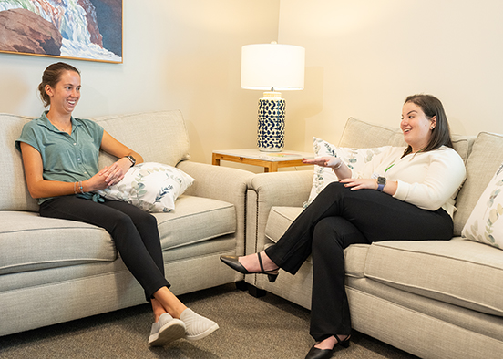 Two people seated on sofas in a counseling room, talking near a lamp and decorative pillows.