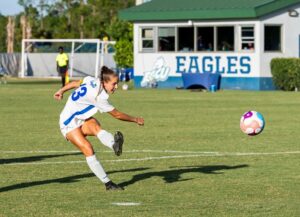 Soccer player kicks ball on a grassy field near a building labeled Eagles