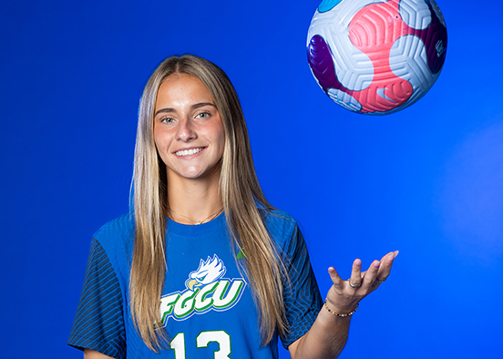 Woman in FGCU jersey tossing a colorful soccer ball against a blue background