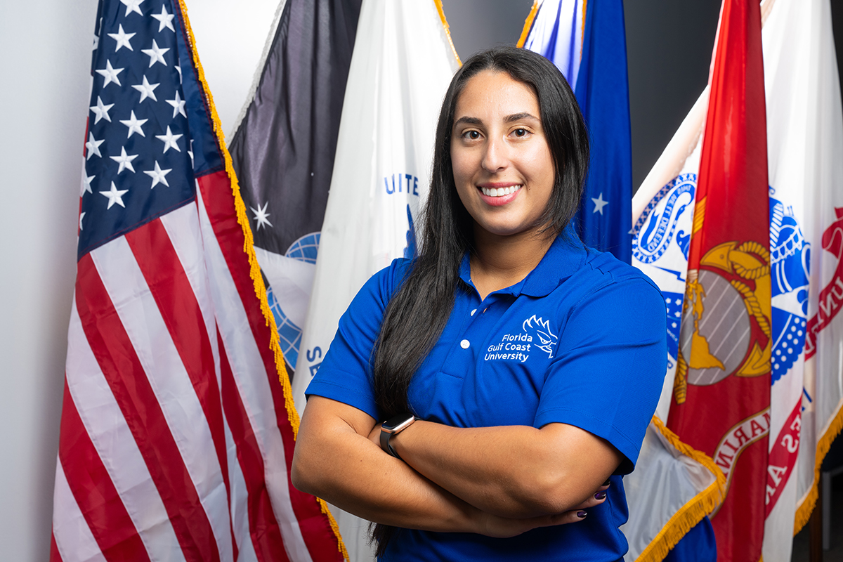 Person in FGCU polo stands with arms crossed in front of U.S. and military service flags