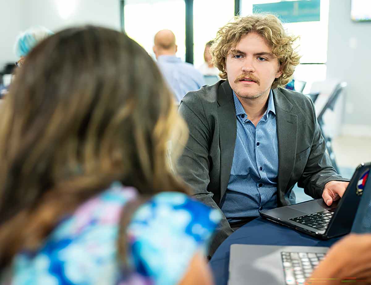 Two people sit at a table with laptops during a meeting in a bright room with large windows
