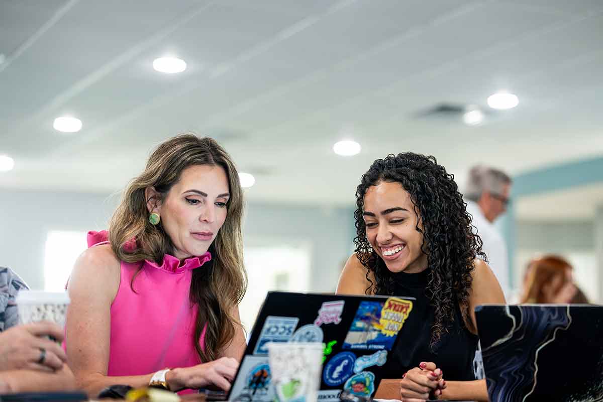 Two women sit at a table with a laptop covered in colorful stickers, appearing to collaborate in a bright room with overhead lights and windows in the background