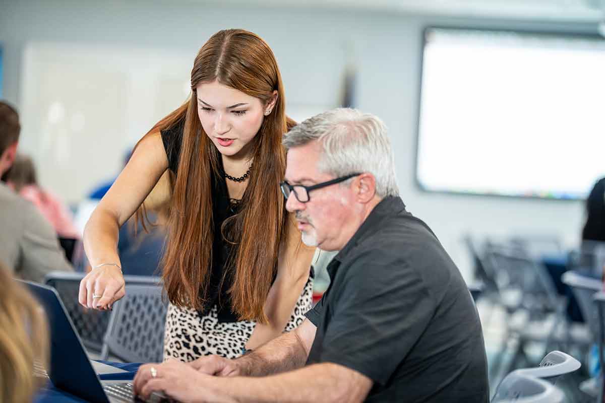 A young woman stands and points at a laptop screen while a middle-aged man in a grey polo shirt and glasses sits and works on the laptop in a meeting space with rows of chairs