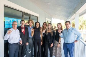 A group of eight people standing on an outdoor covered walkway in front of a building, dressed in business or semi-formal attire, with everyone making the FGCU's Wings Up hand gesture. A sign on the glass door reads “Sanibel & Captiva Islands Association”