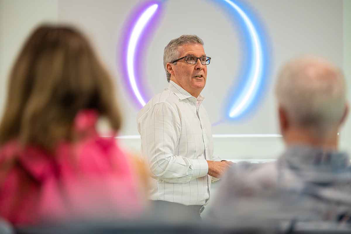 A man wearing a white, long-sleeve shirt stands and speaks in front of a wall with a glowing circular design, while two seated individuals in the foreground listen