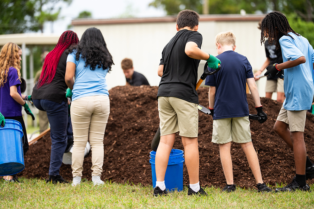Group of students working together to move soil with buckets and shovels near a large mulch pile.