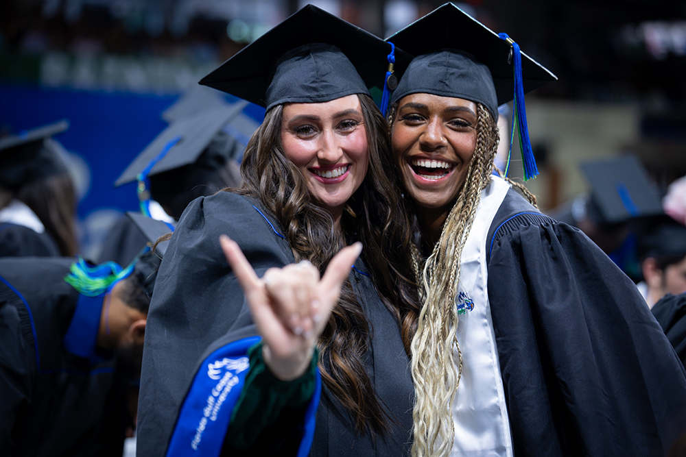 Graduates in caps and gowns pose together, one making a hand gesture with extended pinky and index fingers.
