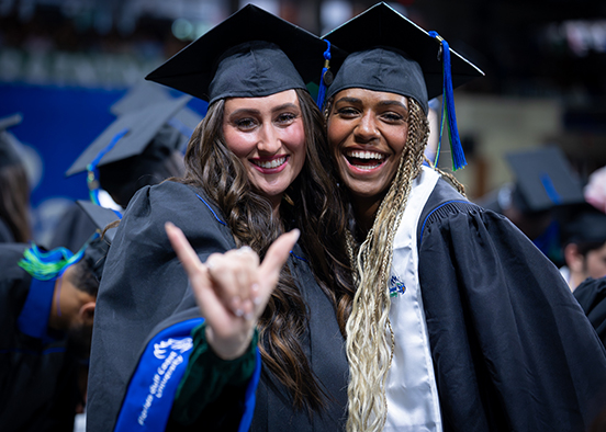 Graduates in caps and gowns pose together, one making a hand gesture with extended pinky and index fingers.