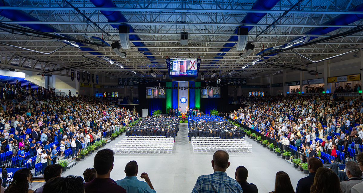 Crowd-filled arena with graduates in caps and gowns seated in rows facing a stage with banners and screens.