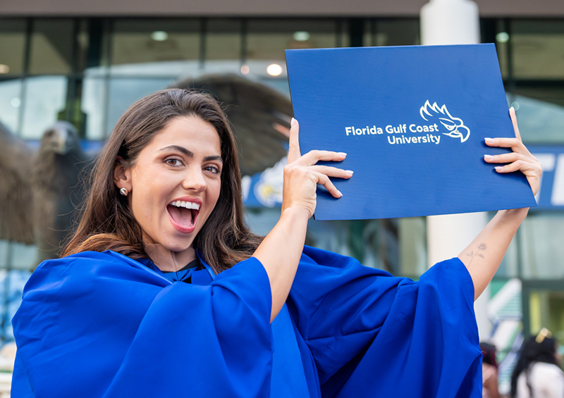 Person in a blue graduation gown holds up a Florida Gulf Coast University diploma folder outdoors.