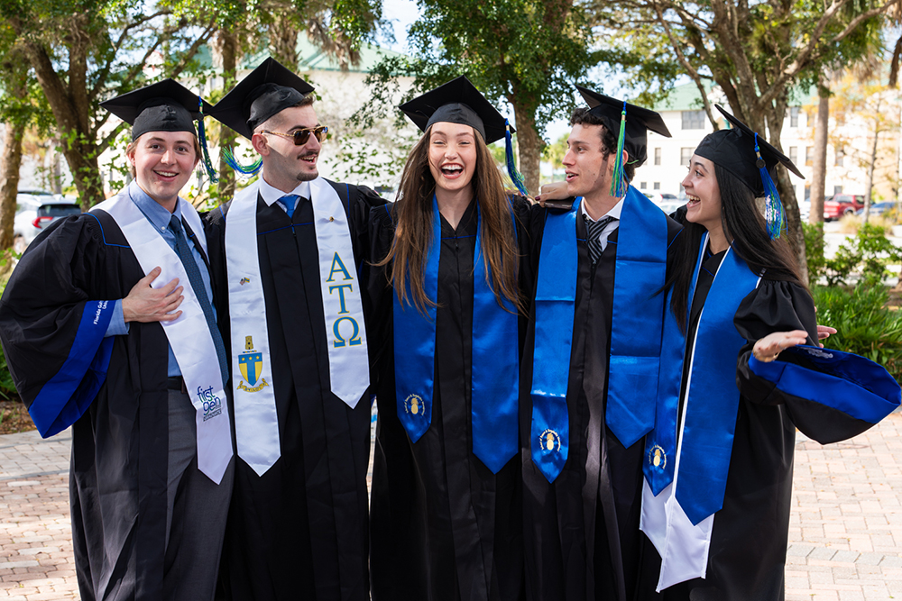 Five graduates in caps and gowns stand together outdoors, wearing blue stoles and white honor sashes.