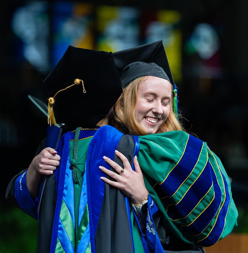 Two people in academic regalia embrace during a graduation ceremony, one holding a diploma in hand.