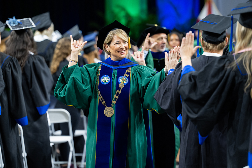 Person in green and blue academic regalia with a medallion raises hands in a celebratory gesture among graduates.