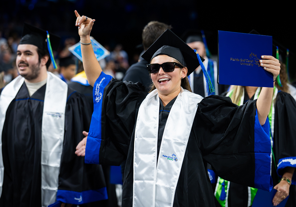 Graduate in cap and gown raises one arm and holds an FGCU diploma folder during commencement ceremony.